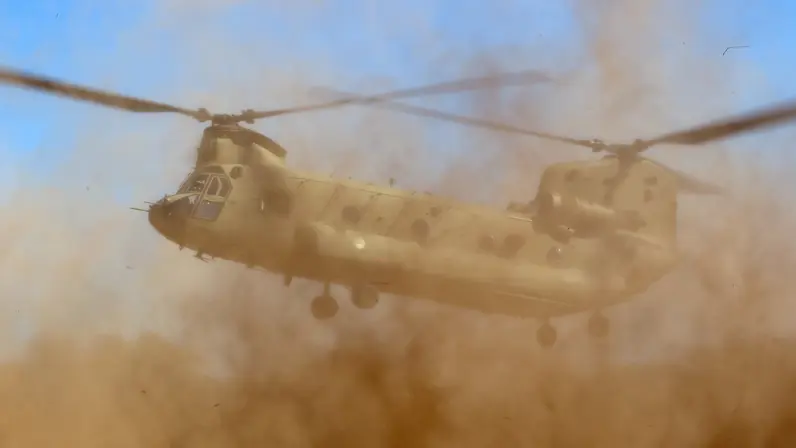 Een militaire helikopter vliegt laag boven een stoffige grond, met zand en stof die opwaaien door de rotorbladen.