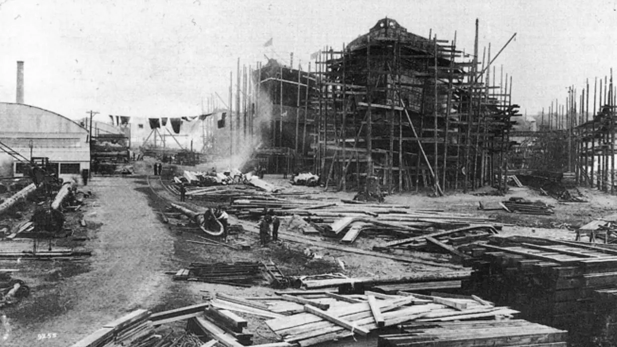 Een drukke bouwplaats met hout, stellingen, een half afgebouwd gebouw en arbeiders in de verte.