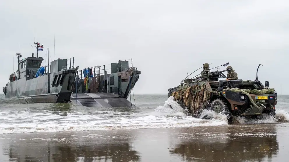 LCU-landingsvaartuig ligt vlakbij het strand, en een Fennek-verkenningsvoertuig met 2 militairen erin rijdt op de voorgrond. 