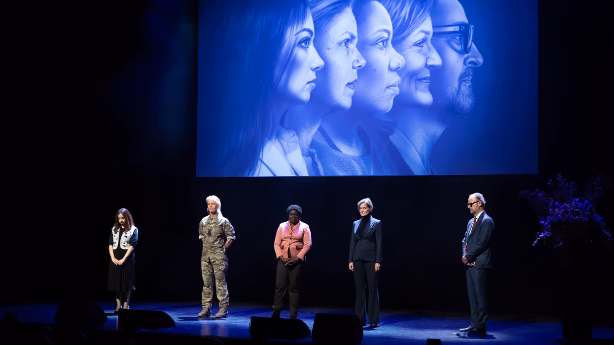 De deelnemers aan de podcast staan op een podium. Achter hen zijn hun gezichten te zien op een scherm. 