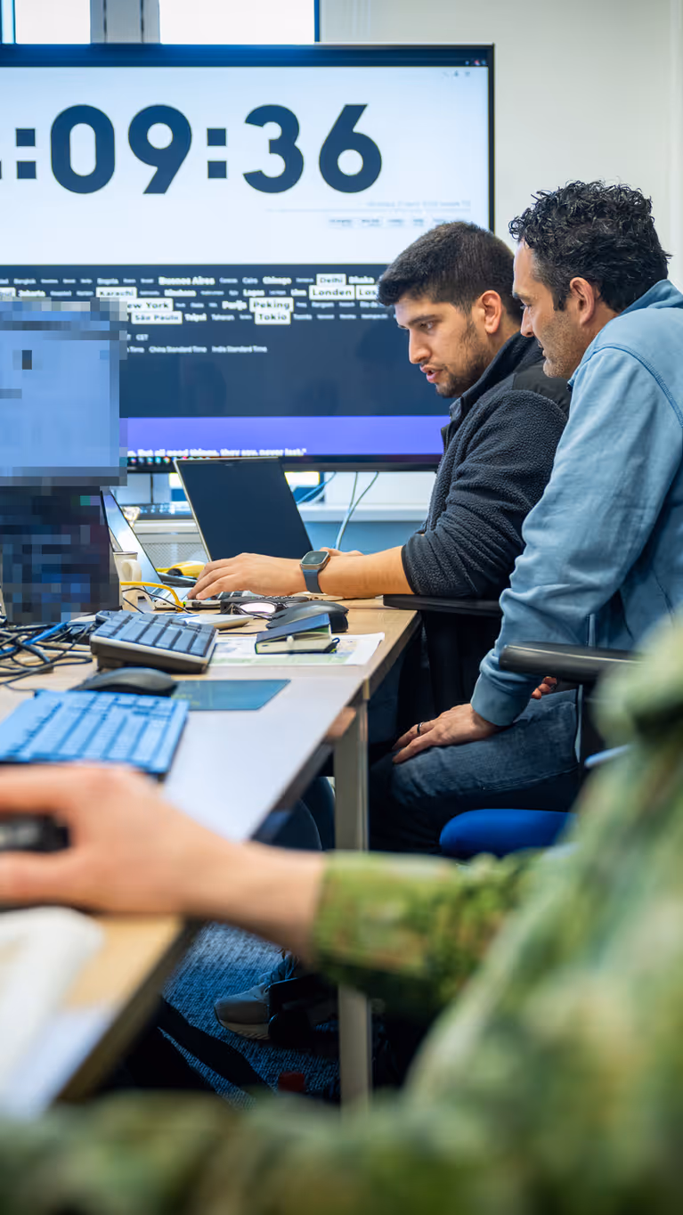 Drie mannen, van wie een Nederlandse militair, zitten achter hun PC aan een bureau met op de achtergrond een groot computerscherm. 