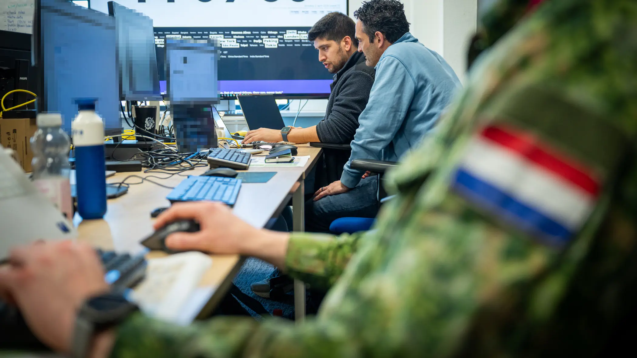 Drie mannen, van wie een Nederlandse militair, zitten achter hun PC aan een bureau met op de achtergrond een groot computerscherm. 