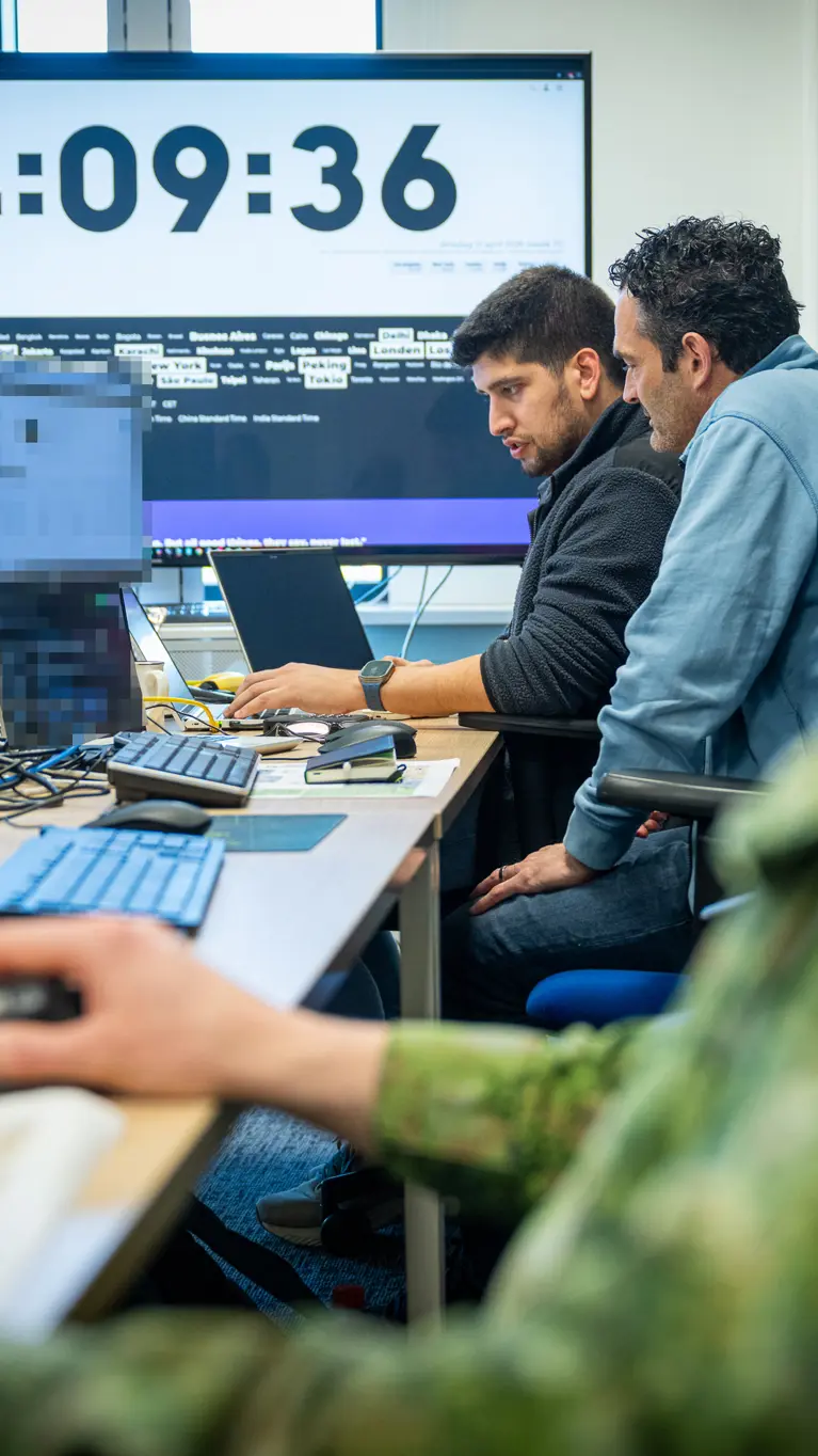 Drie mannen, van wie een Nederlandse militair, zitten achter hun PC aan een bureau met op de achtergrond een groot computerscherm. 