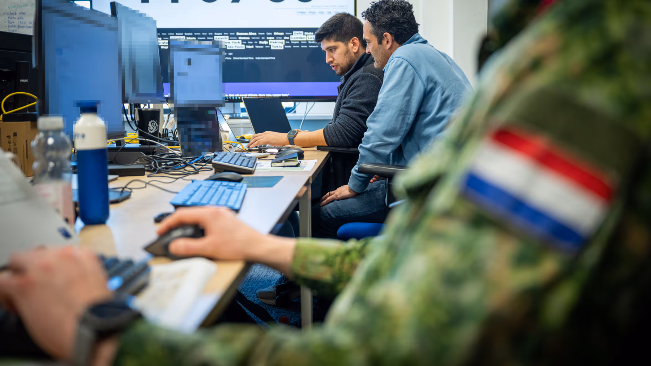 Drie mannen, van wie een Nederlandse militair, zitten achter hun PC aan een bureau met op de achtergrond een groot computerscherm. 
