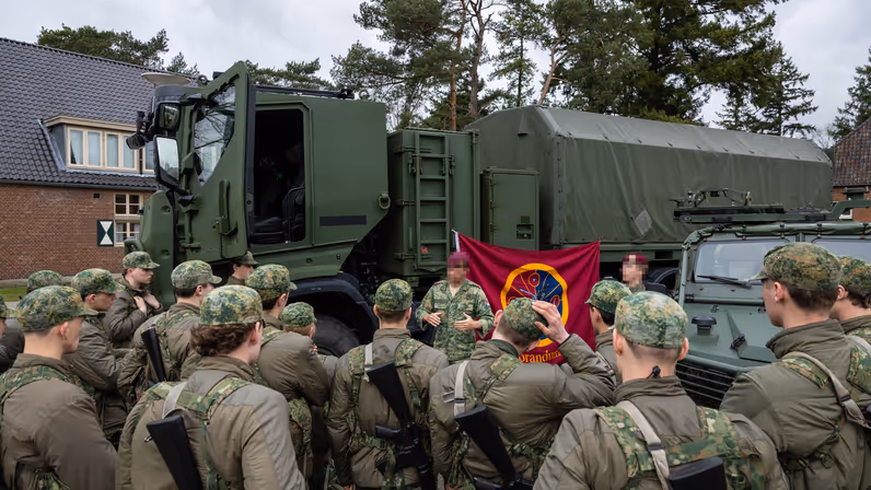  Een groep VeVa-studenten in camouflageuniformen staat in een halve cirkel. Een militair met rode baret geeft uitleg bij twee militaire voertuigen, met een rode vlag met een embleem.