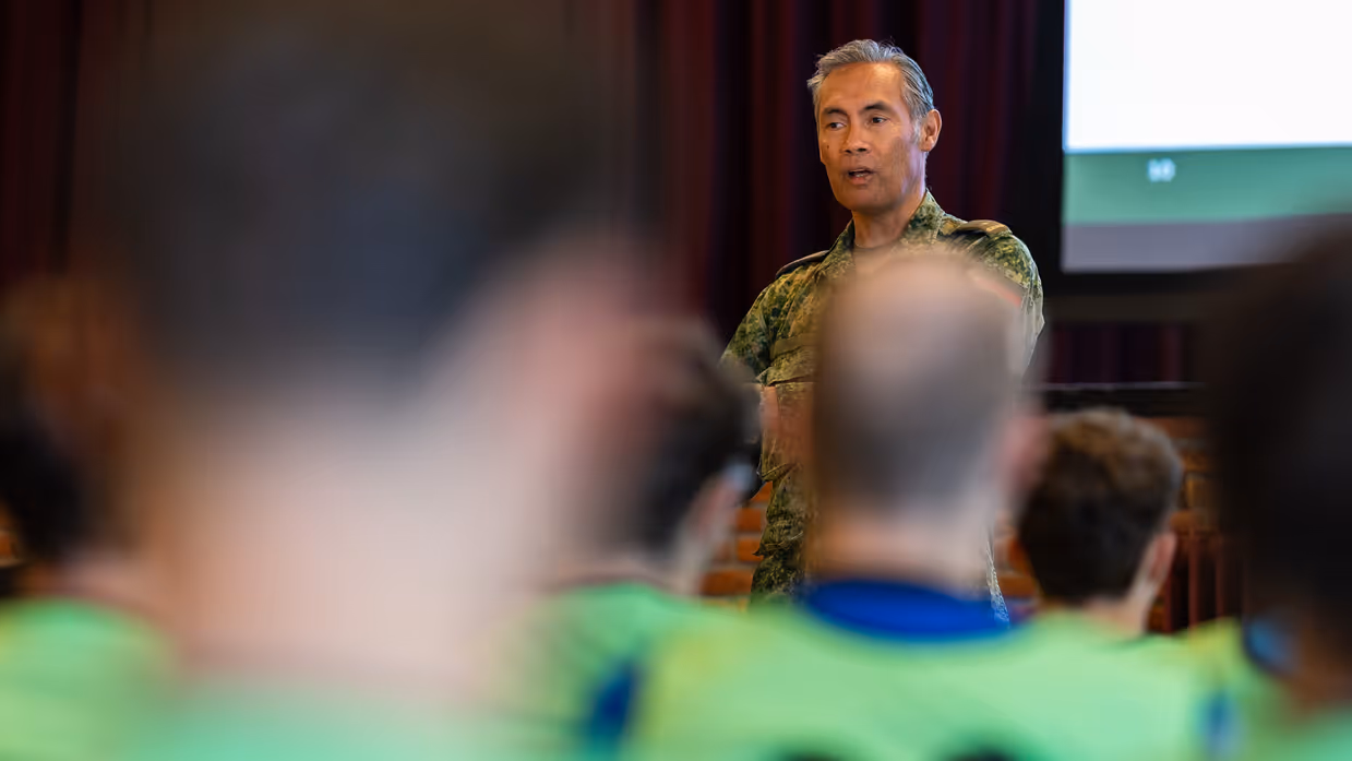 Een groep jongeren in gele vesten luistert in een zaal naar een militair die een presentatie geeft over organisatie en verschillende logo’s op een groot scherm.