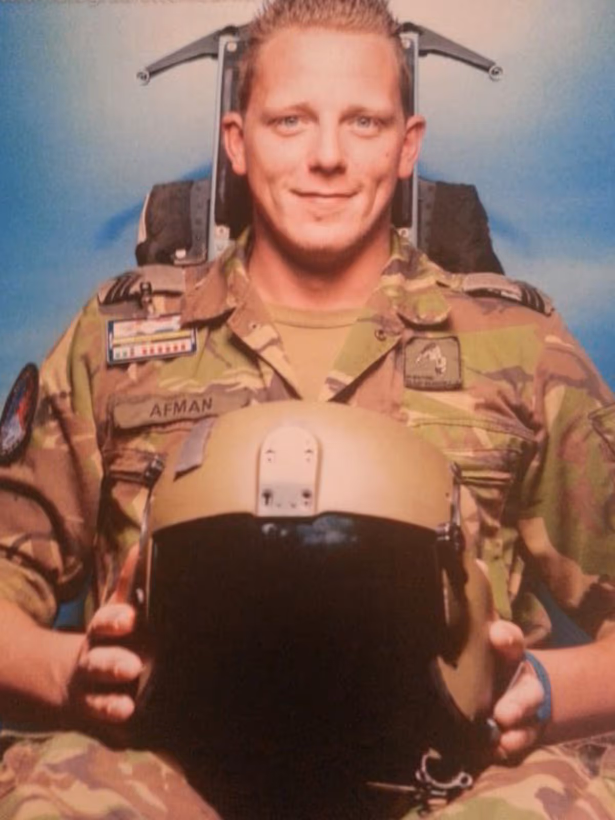 Sergeant Glenn Afman in vlekkenpak op een schietstoel met een vliegerhelm op zijn schoot.