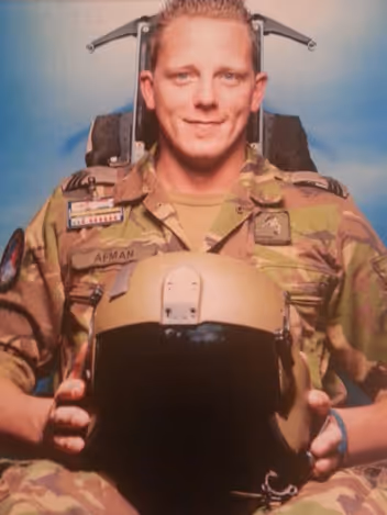 Sergeant Glenn Afman in vlekkenpak op een schietstoel met een vliegerhelm op zijn schoot.