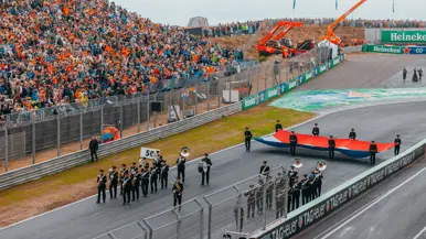 Het Orkest Koninklijke Luchtmacht speelt muziek midden op de racebaan. Achter het orkest houden acht militairen een grote vlag vast die bijna even breed is als het wegdek. Naast de racebaan staat een tribune vol publiek.