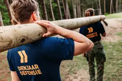 2 jongeren met shirt Dienjaar Defensie sjouwen een boomstam.