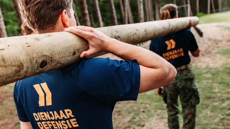 Twee jongeren met shirt Dienjaar Defensie sjouwen een boomstam op de schouders.