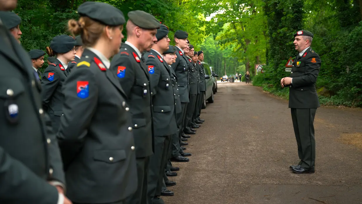Luitenant Kleinbloesem spreekt reservisten toe Twee reservisten lopen met een krans 