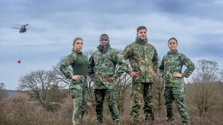 Militairen staan in het veld en dragen een vlekkenpak. Achter hen hangt een helikopter in de lucht.