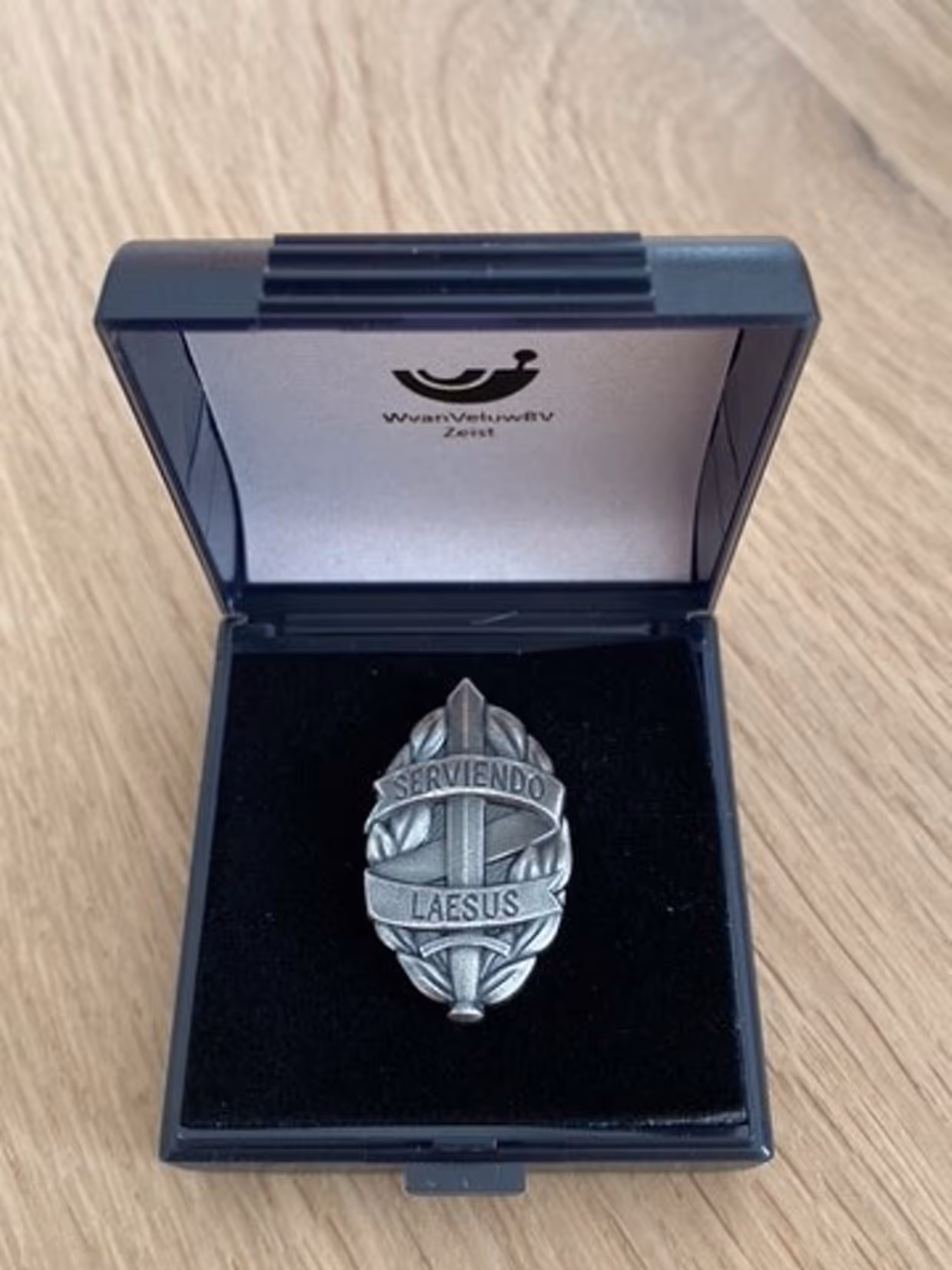 Voorbeeld van een MOD-insigne in een doosje. Het insigne bestaat uit een zwaard van mat witmetaal, omgeven door een banderol met de Latijnse tekst Serviendo Laesus.