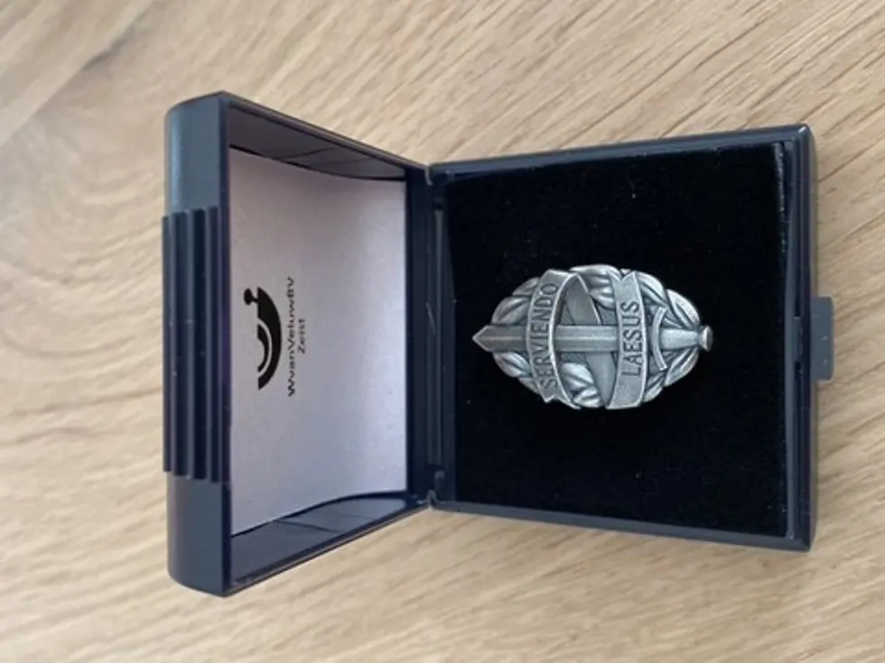 Voorbeeld van een MOD-insigne in een doosje. Het insigne bestaat uit een zwaard van mat witmetaal, omgeven door een banderol met de Latijnse tekst Serviendo Laesus.