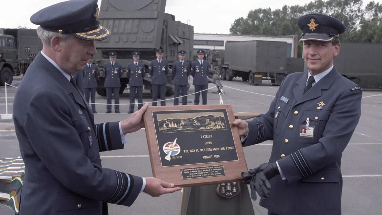 Twee militairen van de Koninklijke Luchtmacht overhandigen elkaar een plaquette tijdens de ceremonie voor de ingebruikname van Patriot in 1986. Op de achtergrond staat een Patriot-lanceerinrichting opgesteld.