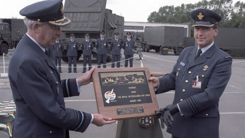 Twee militairen van de Koninklijke Luchtmacht overhandigen elkaar een plaquette tijdens de ceremonie voor de ingebruikname van Patriot in 1986. Op de achtergrond staat een Patriot-lanceerinrichting opgesteld.