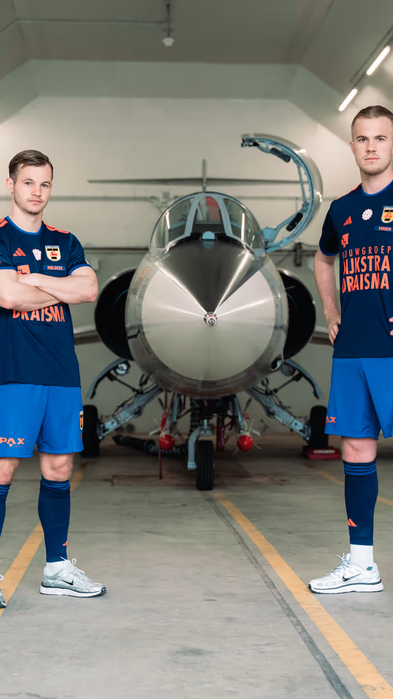 Twee mannen staan in een voetbaltenue naast een oude straaljager in een zaal.