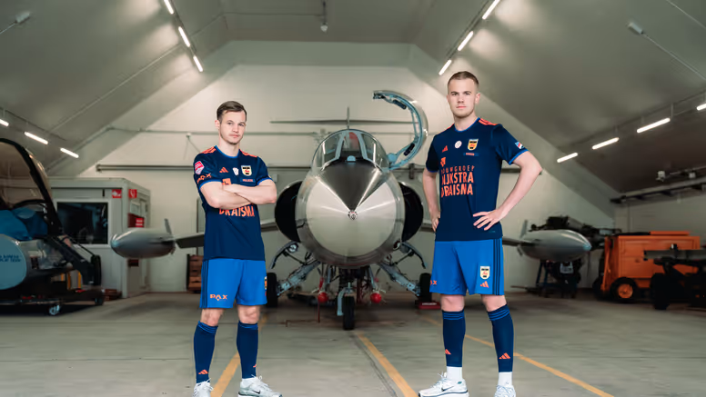 Twee mannen staan in een voetbaltenue naast een oude straaljager in een zaal.