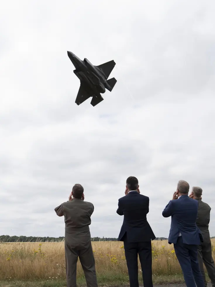 Vier mannen op de rug bekeken die hun oren dichthouden voor de laag overvliegende F-35.