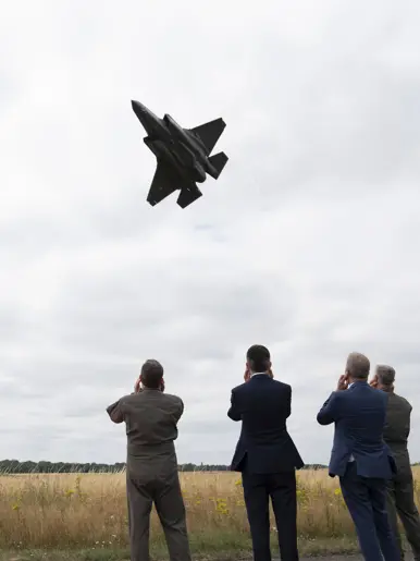 Vier mannen op de rug bekeken die hun oren dichthouden voor de laag overvliegende F-35.