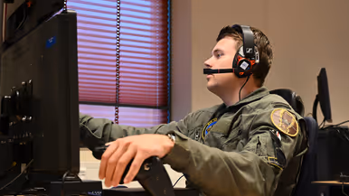 Vlieger met headset op achter een bureau met computerscherm, zijn linkerhand rust op een zwarte hendel.