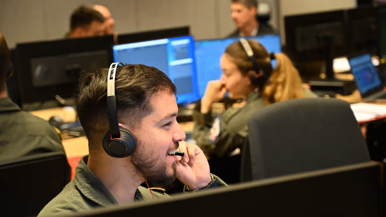 : Militair met headset op zit achter een beeldscherm en spreekt lachend door zijn microfoon. Op de achtergrond meerdere militairen aan bureaus met schermen.
