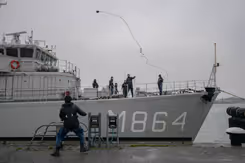 Marineschip legt aan bij kade. Militair gooit touw naar collega op kade.