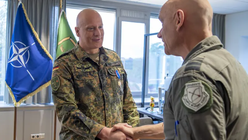 Steur schudt Gerhartz de hand in een kantoor met op de achtergrond onder meer de NAVO-vlag. Gerhartz draagt de medaille Ereteken voor Verdienste in Goud. 
