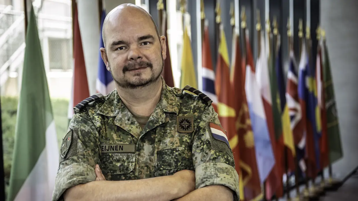 Portret van een militair met armen over elkaar. Achter hem staat een rij vlaggen.