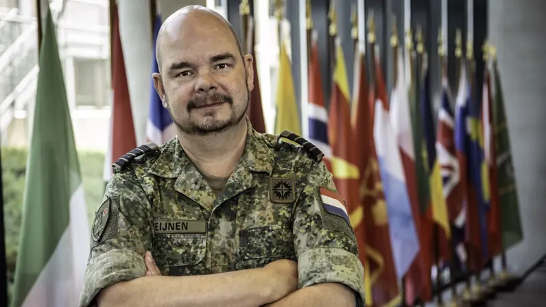 Portret van een militair met armen over elkaar. Achter hem staat een rij vlaggen.