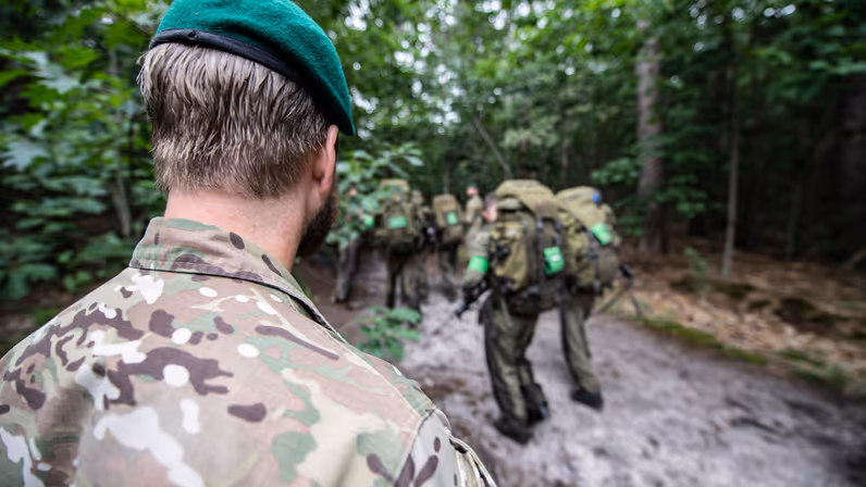 Een instructeur met groene baret kijkt hoe cursisten zich tijdens de commando-opleiding in groene overals verplaatsen.