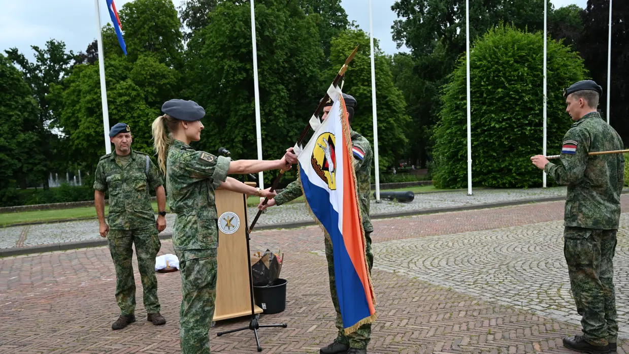 Duijnhouwer overhandigt aan Schilder een vlag op een buitenterrein. Erachter en ernaast staan nog twee militairen. Allen zijn gekleed in gevechtstenue.