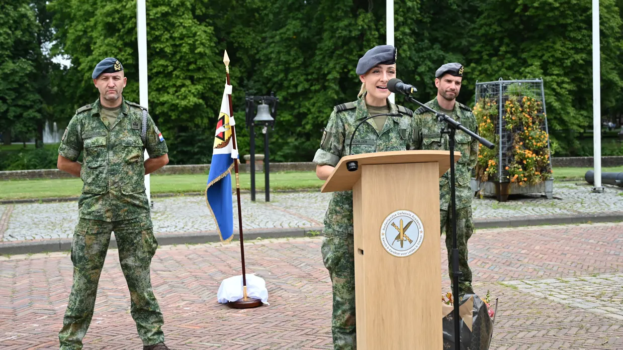 Schilder spreekt vanachter een spreekgestoelte op een buitenterrein. Op de achtergrond staan Duijnhouwer en nog een militair. Allen zijn gekleed in gevechtstenue.