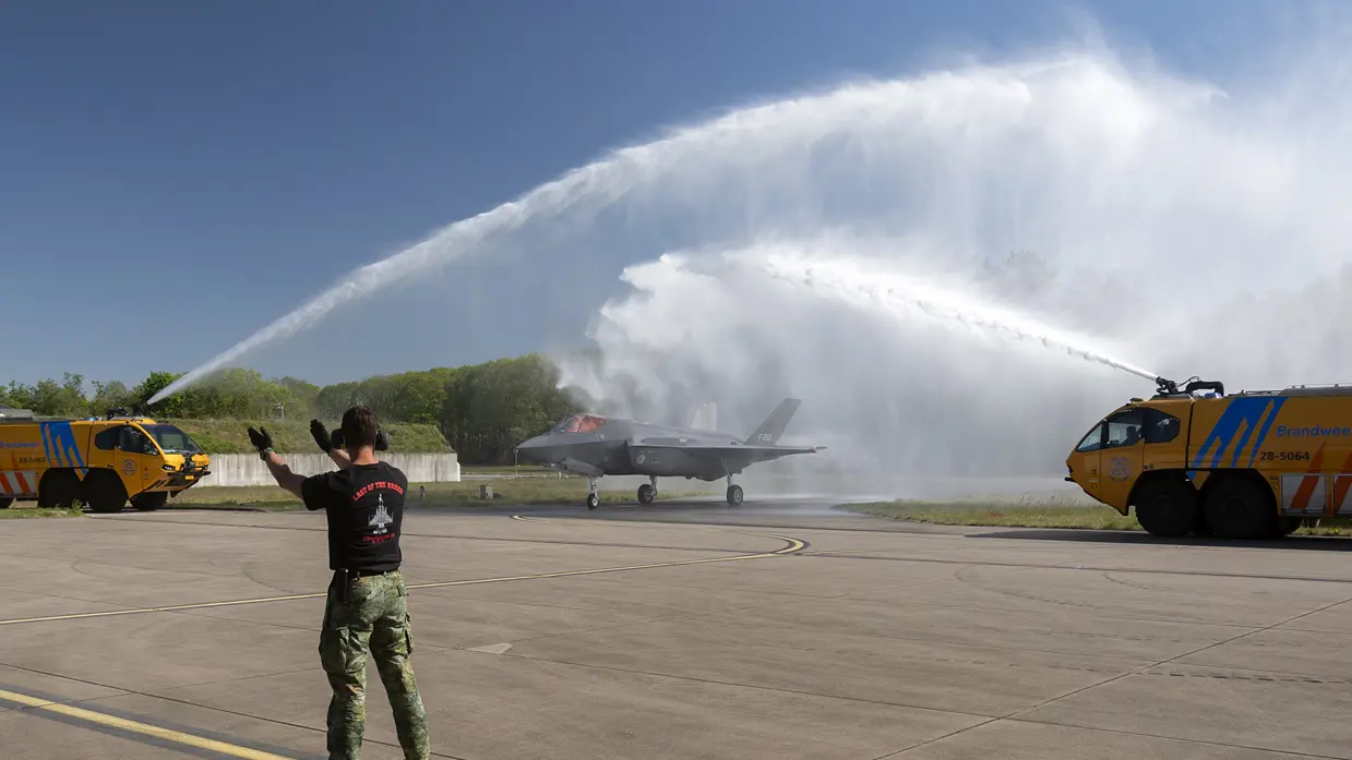 Twee gele brandweerauto’s spuiten van weerszijden een waterboog over de F-35 met staartnummer F-052.