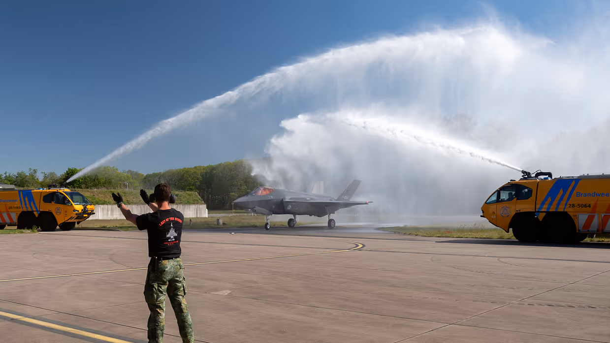 Twee gele brandweerauto’s spuiten van weerszijden een waterboog over de F-35 met staartnummer F-052.