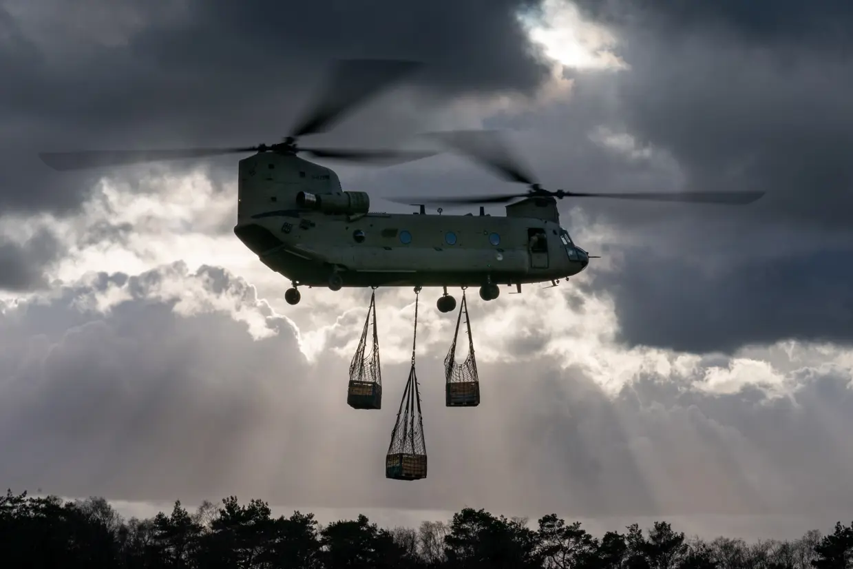 Een Chinook-transporthelikopter in de lucht, er hangen 3 netten met houten kisten aan de onderkant.