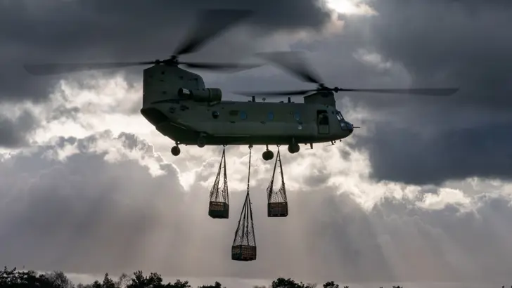 Een Chinook-transporthelikopter in de lucht, er hangen 3 netten met houten kisten aan de onderkant.