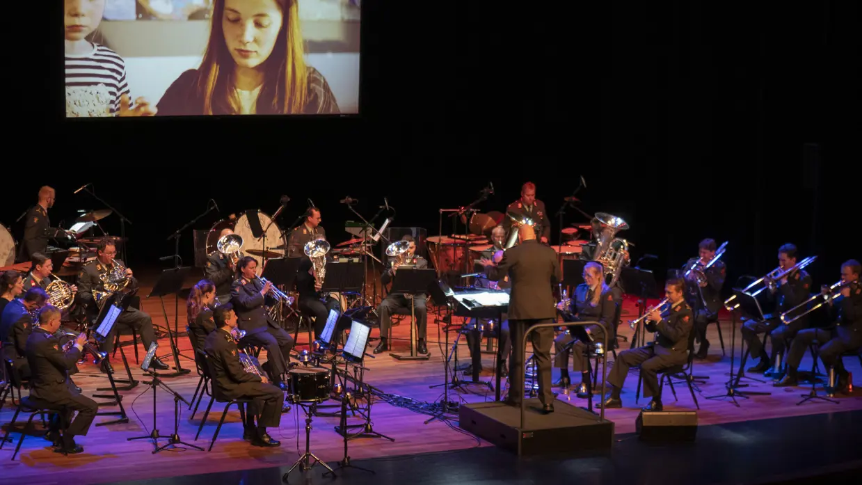 Een militaire orkest speelt muziek met op de achtergrond een beeldscherm. Daarop een mevrouw en een kind.
