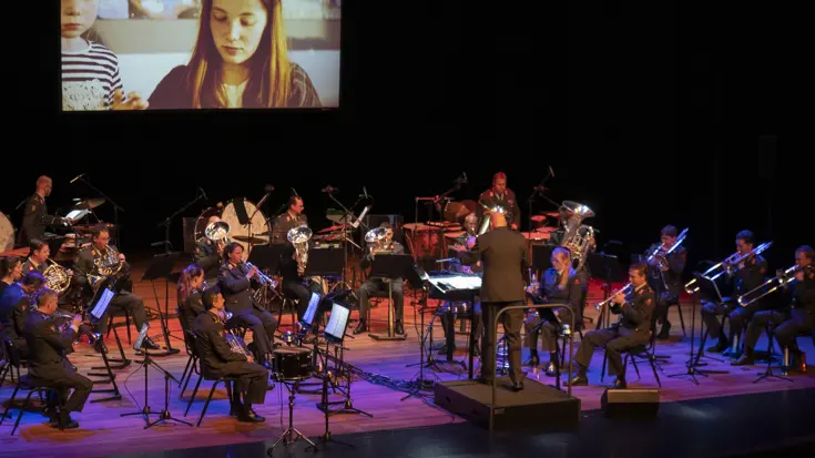 Een militaire orkest speelt muziek met op de achtergrond een beeldscherm. Daarop een mevrouw en een kind.