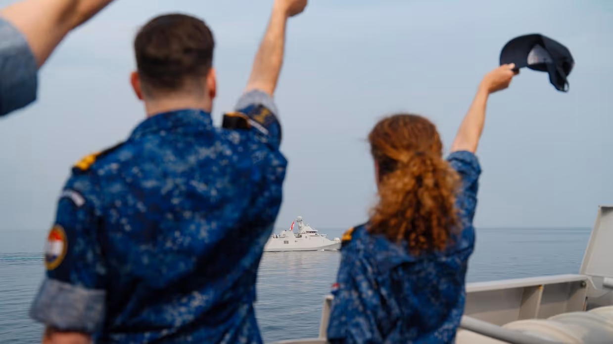 Bemanningsleden van de De Ruyter zwaaien met hun hoofddeksel naar de opvarenden van een Indonesisch fregat dat wegdraait.