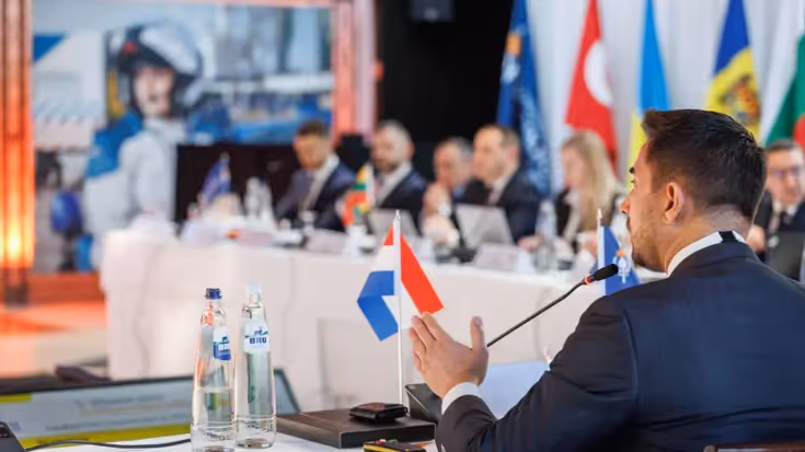 Een Nederlandse afgevaardigde spreek zijn collega’s toe vanachter een tafel met daarop een Nederlands vlaggetje.