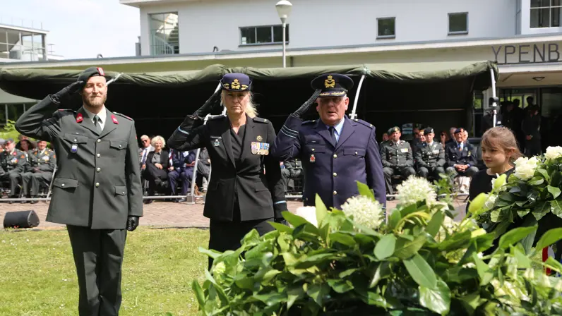 Drie militairen in de houding voor een krans. Achter hen zittend publiek waaronder vele militairen.