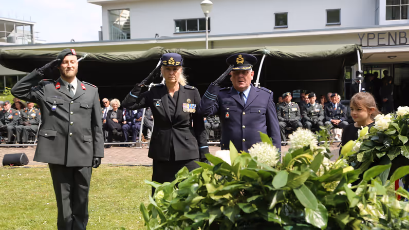 Drie militairen in de houding voor een krans. Achter hen zittend publiek waaronder vele militairen.