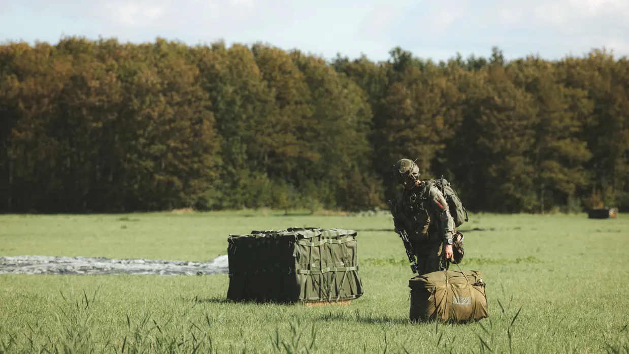 Militair in groen pak met helm, rugtas en wapen heeft groene tas in de hand en staat naast een groen pakket op een pallet. Erachter ligt een parachute in het gras. Op de achtergrond een bosrand.