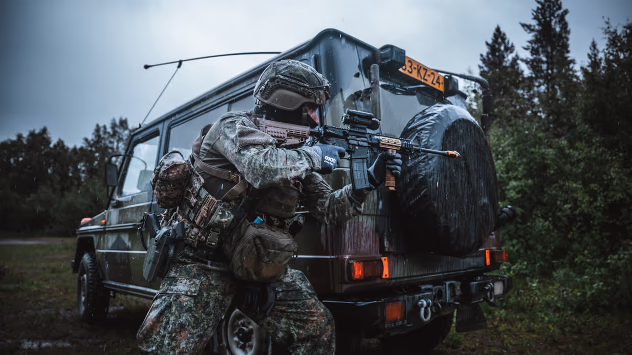 Een gewapende militair met een Colt voor zijn gezicht beweegt langs de achterkant van een groene terreinwagen.