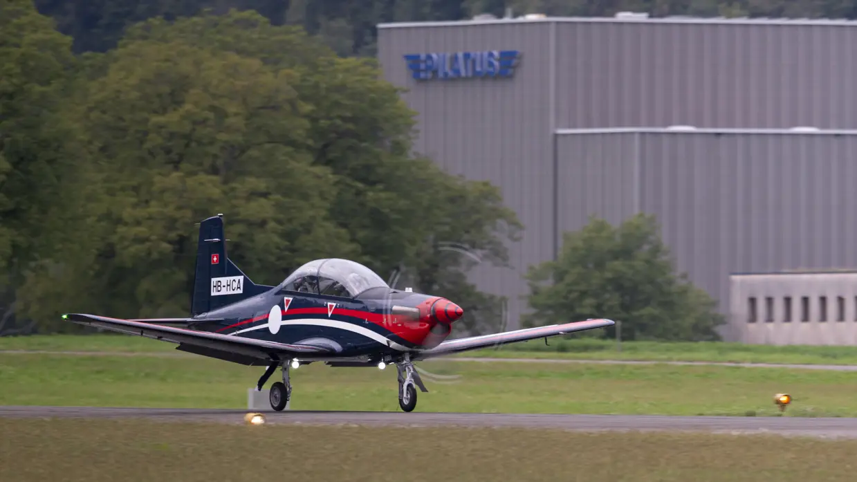 De PC-7 MKX rijdt op hoge snelheid over de startbaan. In de achtergrond een lichtgrijs gebouw met daarop de naam Pilatus.