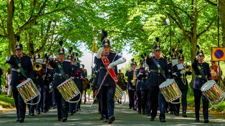 Militaire muzikanten met trommels marcheren onder groene bomen.