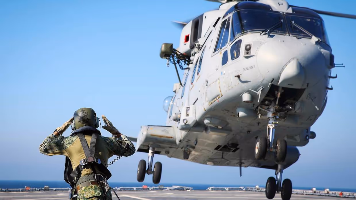 Een lichtgrijze helikopter stijgt op, terwijl een militair op de voorgrond aanwijzingen geeft.