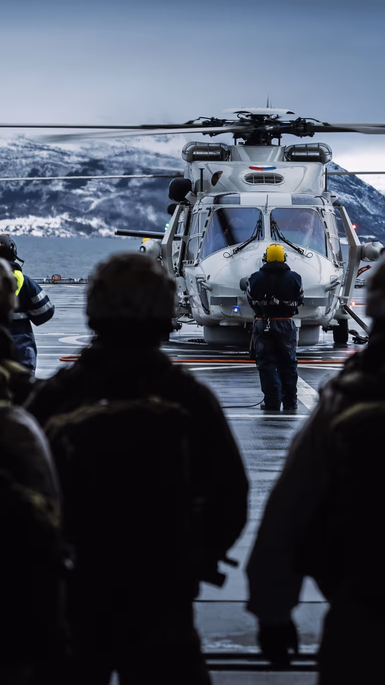 Mariniers, op de rug gezien, wachten op het helidek om in een NH90 te stappen.